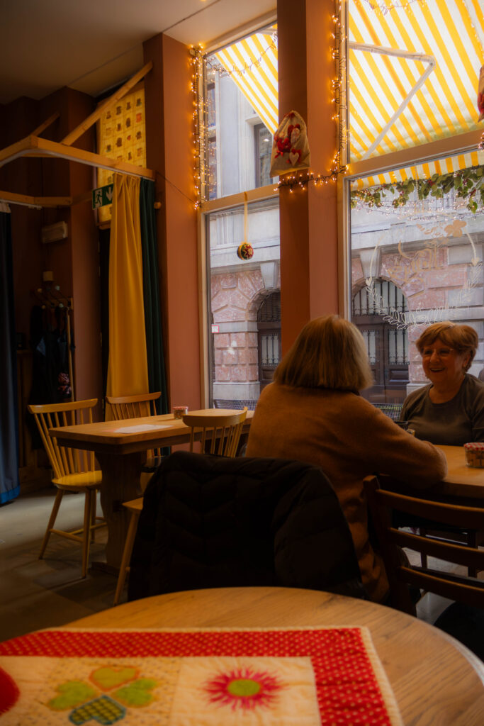 2 dames op leeftijd die aan een tafel zitten in een coffee shop over elkaar en een gesprek hebben. 1van de vrouwen zit met haar rug naar de foto de andere met haar gezicht en deze vrouw lacht een beetje.