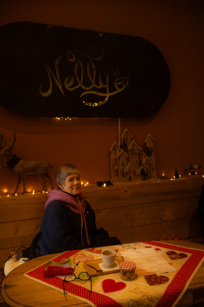 Binnen in een coffee shop aan de muur hangt een groot metaal bord met de naam "Nelly" de naam van de coffee shop. onder het bord is een houten bank en een tafel. er zit ook een dame op de bank onder het bord die een beetje lacht en naar de camera kijkt.