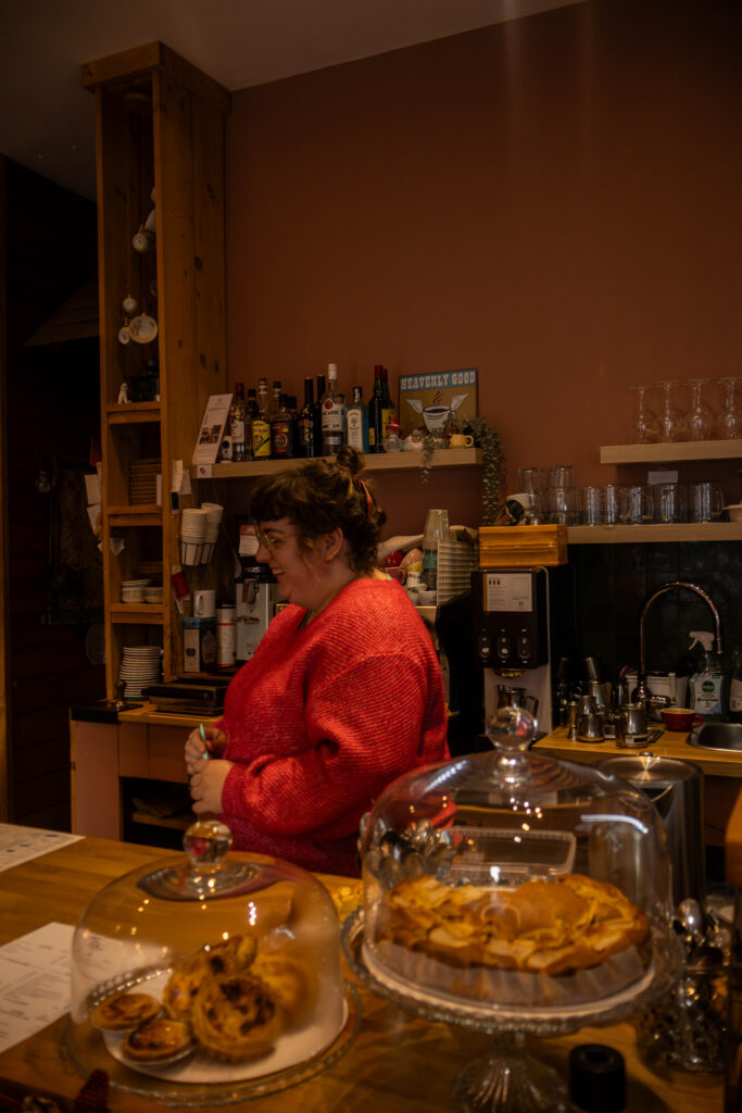 Een foto van een jong volwassen vrouw met bruin haar en een rood / roze trui. Ze staat zijwaarts op de foto en ze kijkt weg maar lacht een beetje. op de voorgrand van de foto staan 2 borden met gebakjes.