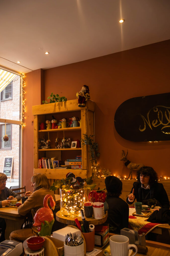 Binnenin de coffee shop "Nelly" op de foto zitten 4 mensen per 2 aan een tafel en ze hebben een gesprek. in de rechter hoek is een stuk van een raam te zien en een kast met verschillende decoratie. Er hangen ook kerst lichtjes op.