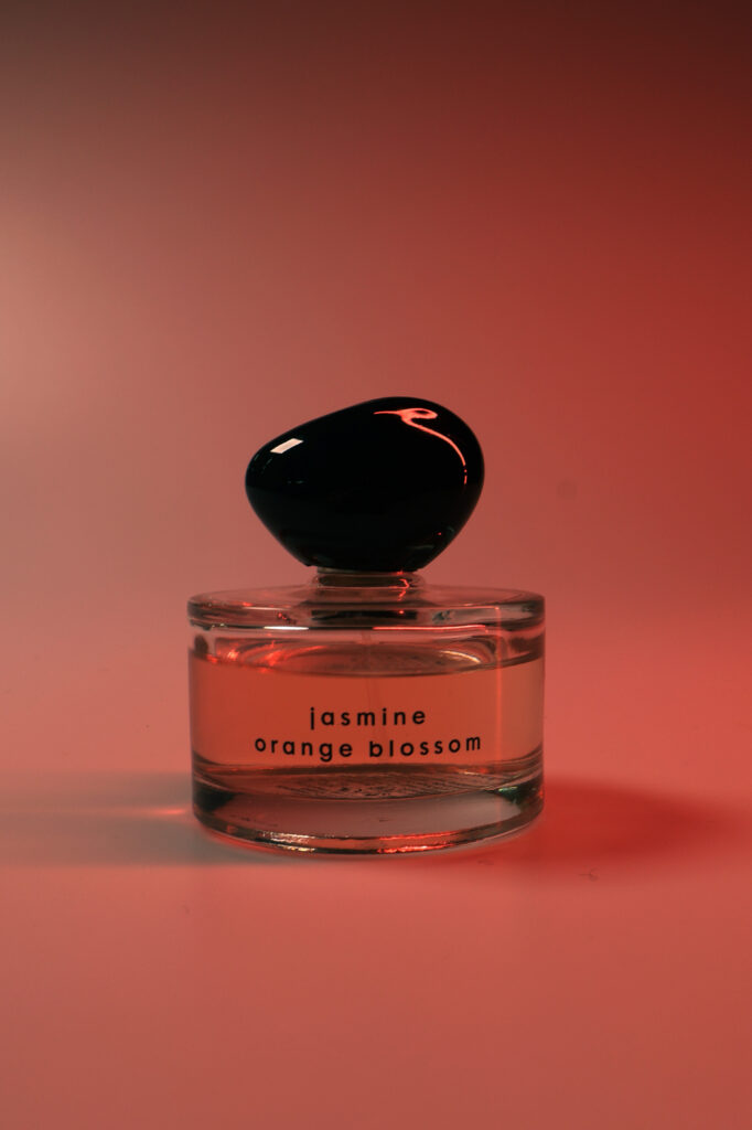 Een glaze doorschijnend parfum flesje met een zwarte asymmetrische dop op het flesje staat een tekst "jasmine orange blossom"