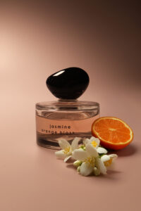 Een glaze doorschijnend parfum flesje met een zwarte asymmetrische dop op het flesje staat een tekst "jasmine orange blossom" Naast het parfum flesje links vanonder licht een halve appelsien en een 4 tal witte bloemetjes.